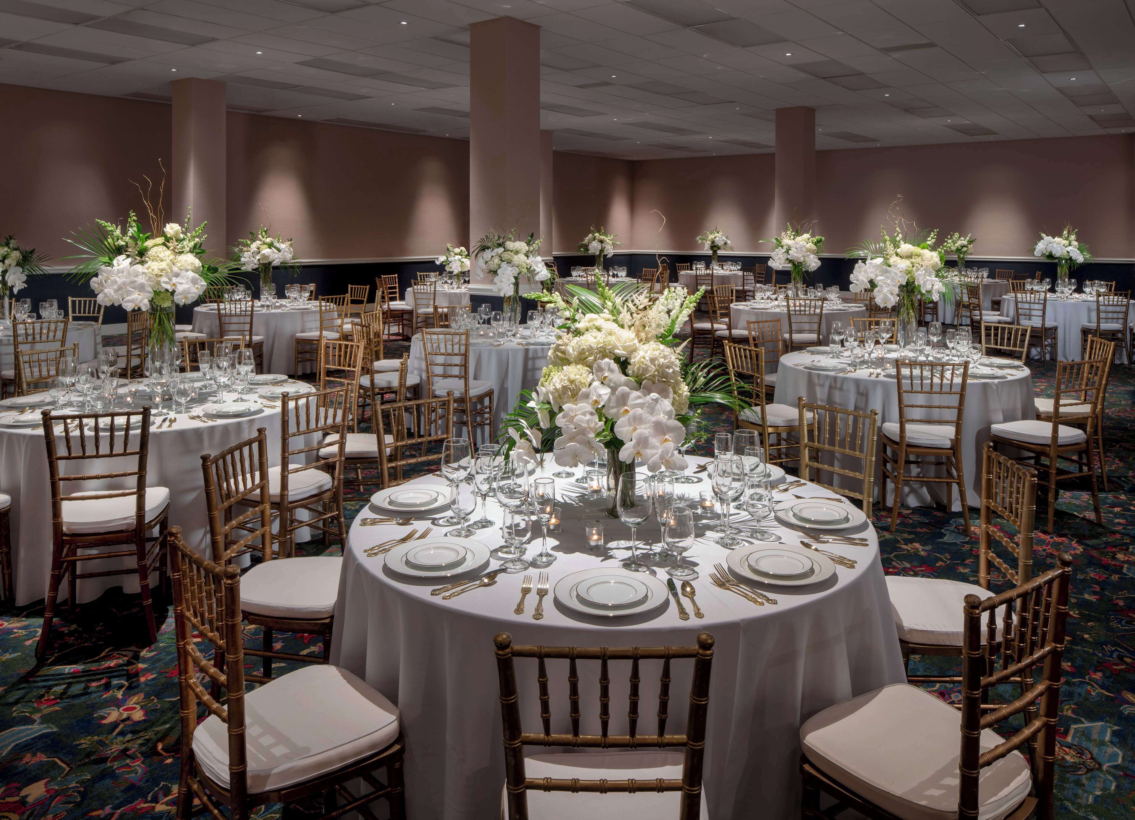 A banquet hall with round tables set for dining, each with white tablecloths, gold chairs, and floral centerpieces.