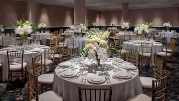 A banquet hall with round tables set for dining, each with white tablecloths, gold chairs, and floral centerpieces.