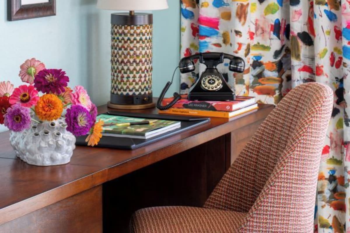 Un pupitre de madera con un florero, una lámpara estampada, un teléfono rotativo y una silla tejida.
