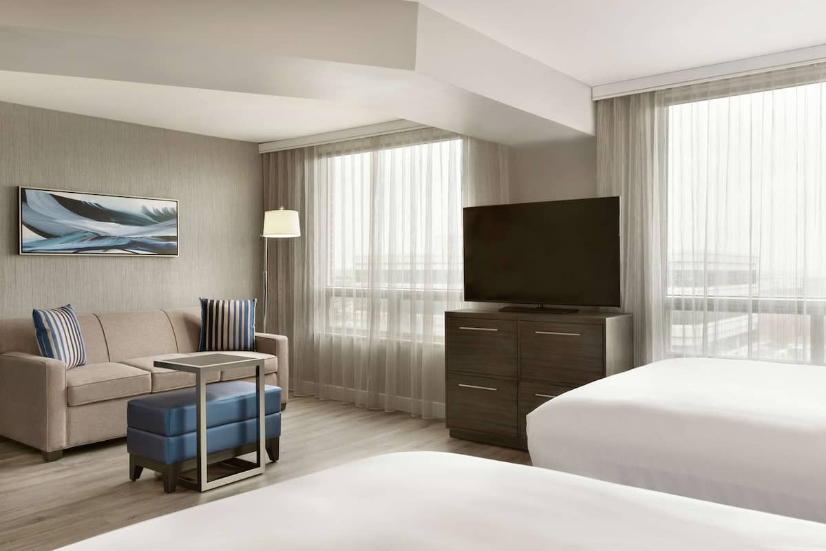 Ein modernes Hotelzimmer mit einem großen Bett, einer Couch, einem kleinen Tisch, einem Fernseher und einem großen Fenster mit transparenten Vorhängen.