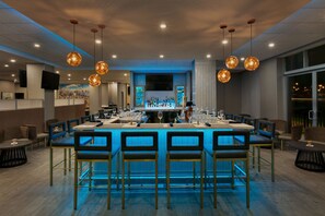 A modern bar area with a long counter, multiple bar stools, and hanging pendant lights.