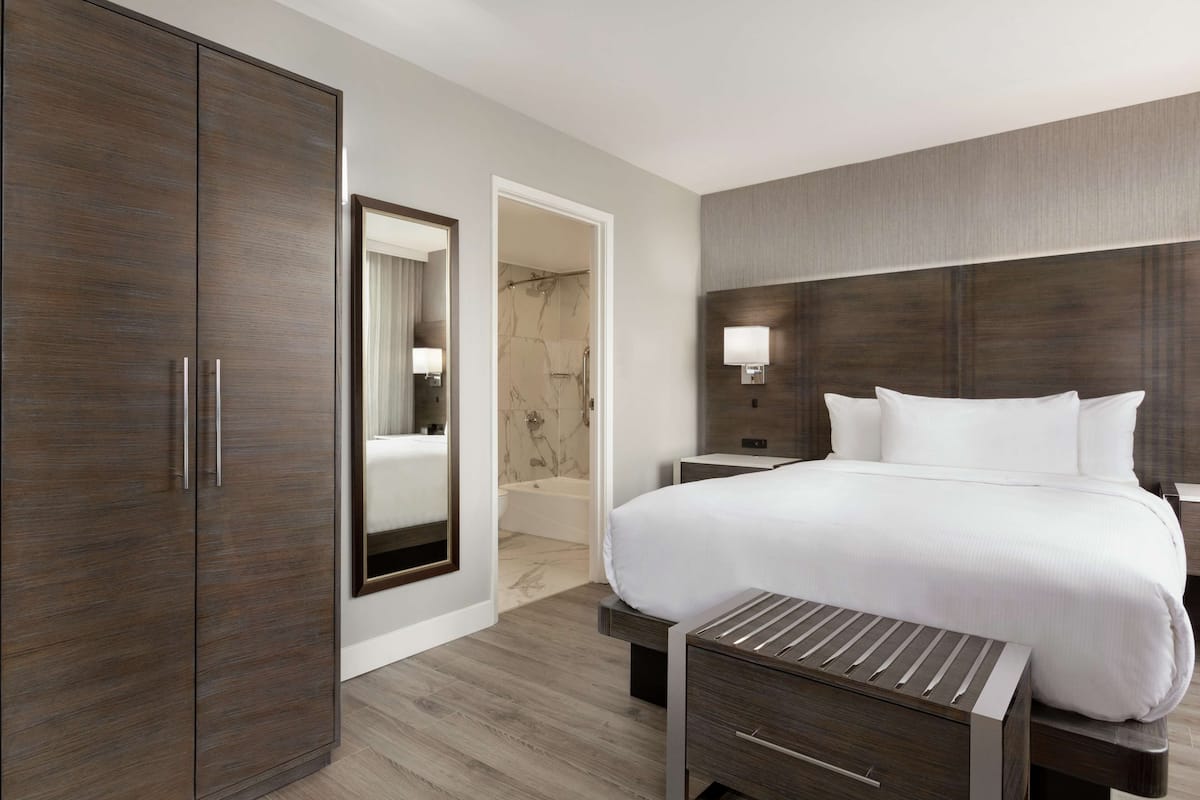 Ein modernes Hotelzimmer mit einem großen Bett, einem Kleiderschrank, einem Badezimmer mit Badewanne und einem Nachttisch mit Lampe.