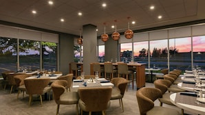 Un restaurant avec des tables et des chaises rondes, de grandes fenêtres offrant une vue sur le coucher du soleil et des luminaires suspendus.