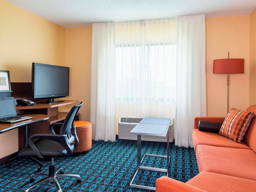 A hotel room with a desk, laptop, television, orange sofa, and a window with curtains.