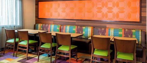 A dining area with a colorful carpet, wooden chairs with green cushions, and a wall with a geometric pattern.