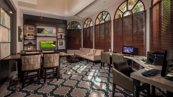 Um hall de hotel com uma televisão grande, assentos confortáveis, uma mesa com computadores e um carpete estampado.