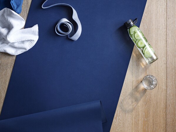 Una colchoneta azul, una toalla blanca, una banda de yoga enrollada, una botella de agua con rodajas de pepino y un vaso de agua sobre una superficie de madera.