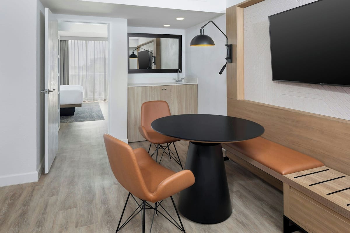 Ein modernes Hotelzimmer mit Essbereich, ausgestattet mit einem runden Tisch, zwei Stühlen, einer Bank, einem Fernseher und einem Spiegel.