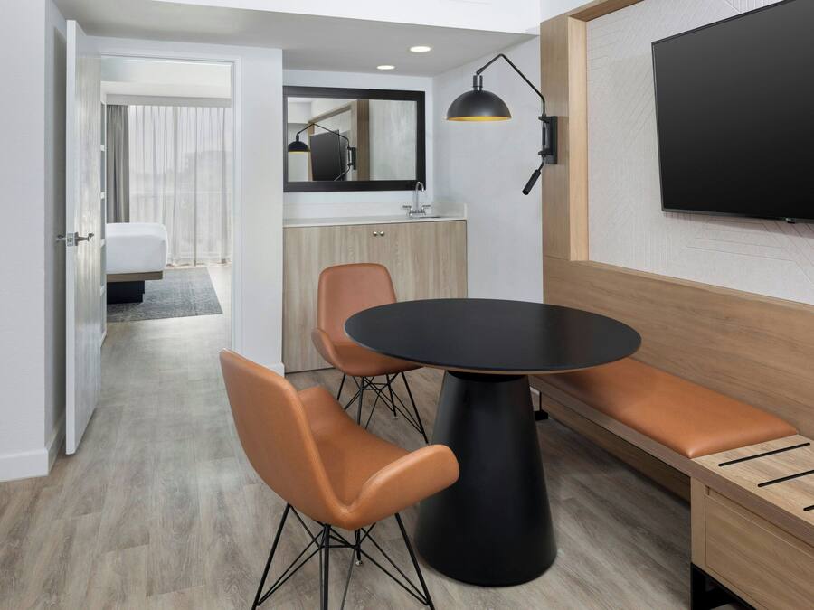 Een moderne hotelkamer met een zithoek, bestaande uit een ronde tafel, twee stoelen, een bank, een televisie en een spiegel.