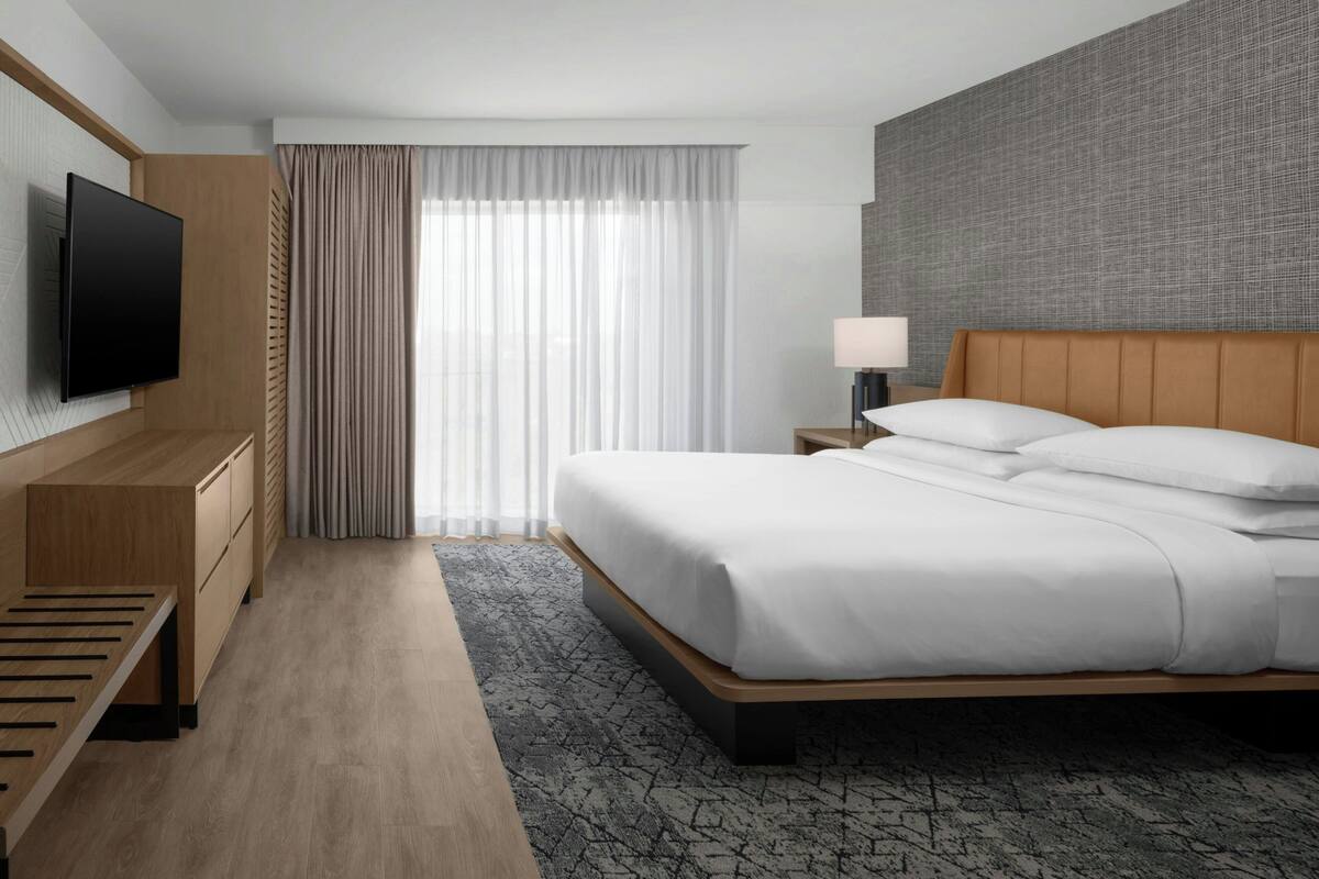 Ein Hotelzimmer mit Bett, Wandfernseher, Schreibtisch, Lampe und einem Fenster mit Vorhängen.