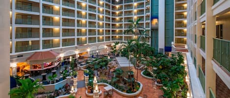 Un vestíbulo de hotel de varios niveles con múltiples balcones, zonas de comedor interiores y un elemento acuático central rodeado de vegetación.