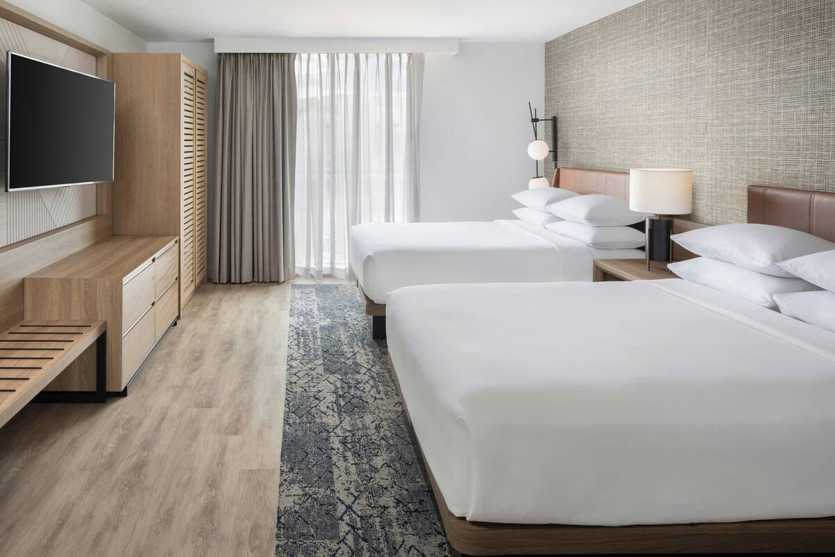 Ein Hotelzimmer mit zwei Betten, einem Fernseher, einem Schreibtisch und einem großen Fenster mit Vorhängen.