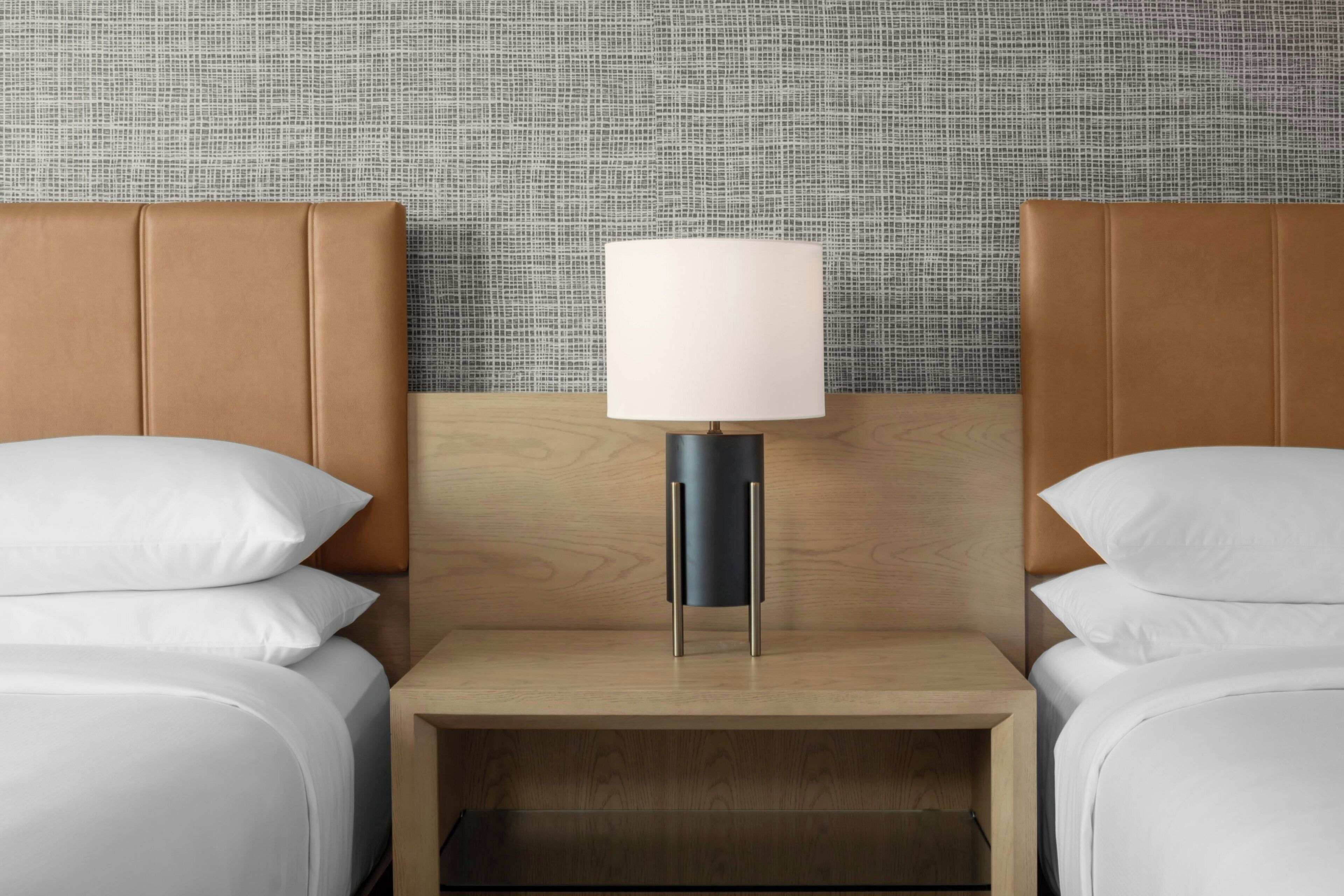 Ein Hotelzimmer mit einem hölzernen Kopfteil, einem Nachttisch mit Lampe und zwei Betten mit weißer Bettwäsche.