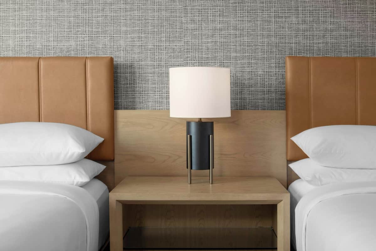 Ein Hotelzimmer mit einem hölzernen Kopfteil, einem Nachttisch mit Lampe und zwei Betten mit weißer Bettwäsche.
