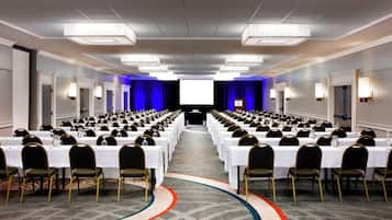 A conference room with rows of tables and chairs, a large screen at the front, and a carpeted floor.