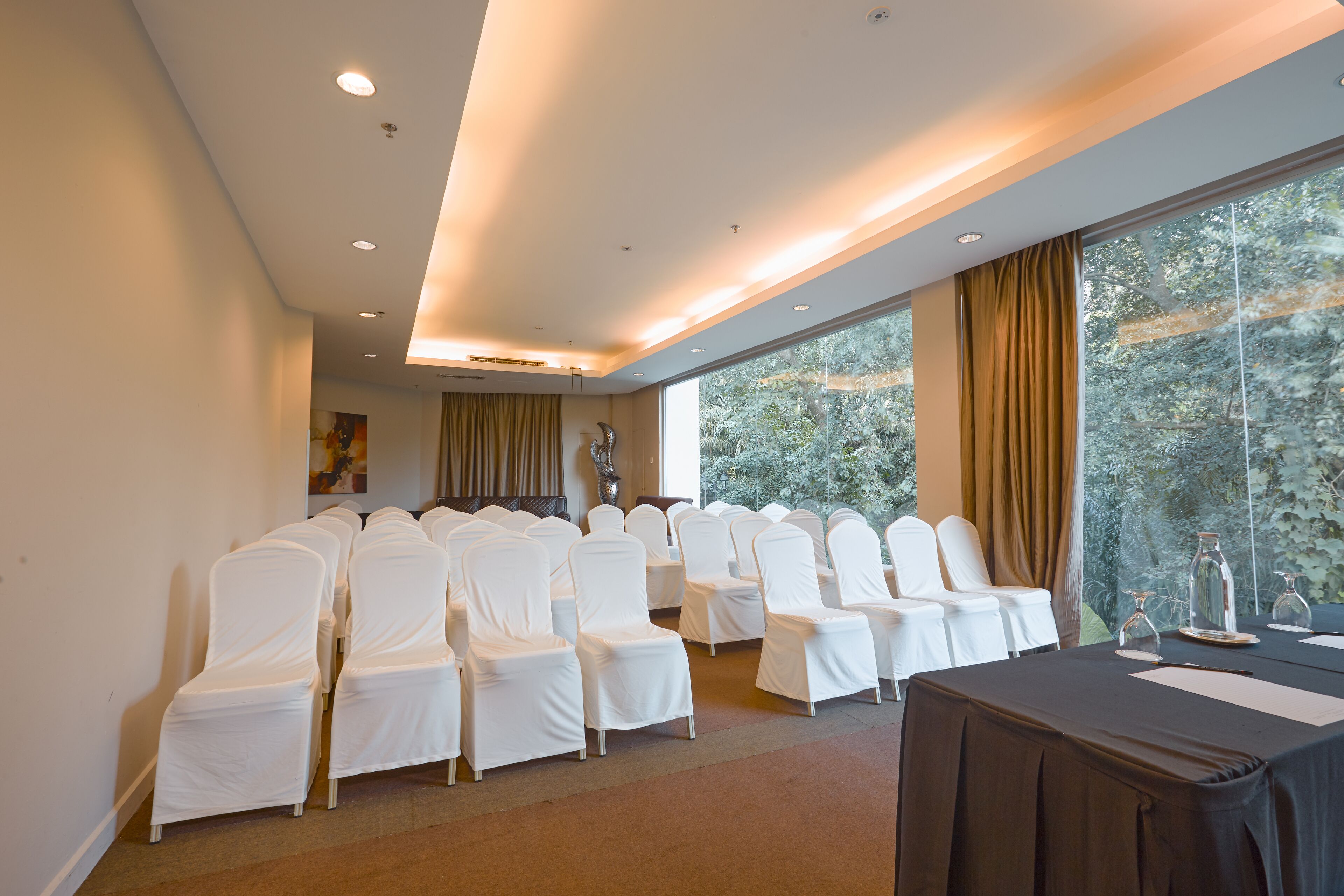 Uma sala de conferências com fileiras de cadeiras cobertas de branco, uma mesa com toalha escura e grandes janelas com cortinas.