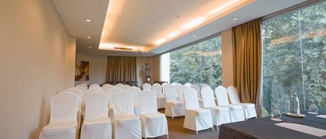 Uma sala de conferências com fileiras de cadeiras cobertas de branco, uma mesa com toalha escura e grandes janelas com cortinas.