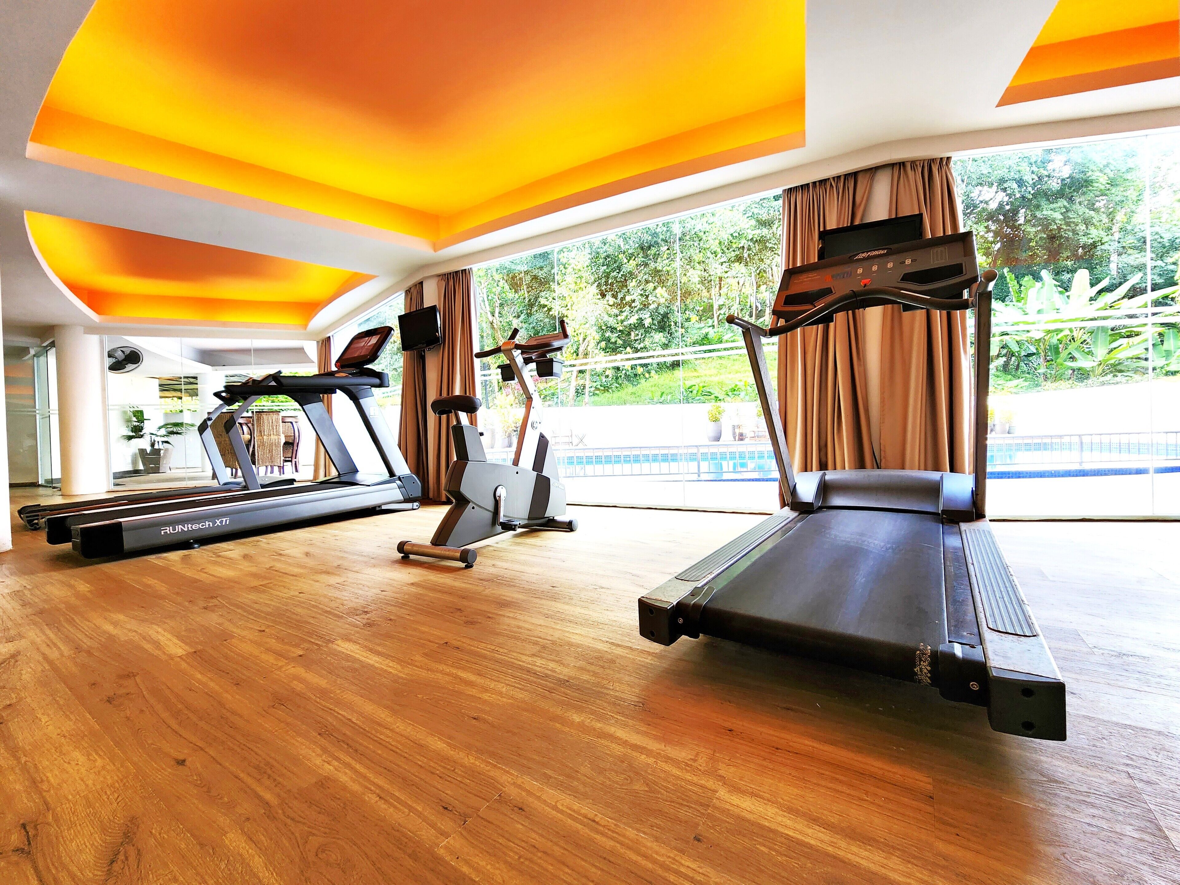 A gym with treadmills and an elliptical machine, large windows with curtains, and a view of greenery outside.
