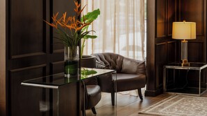 Une chambre d’hôtel moderne avec des lambris en bois foncé, une table basse en verre, un fauteuil en cuir, une petite table d’appoint avec une lampe, et une vue sur l’extérieur à travers des rideaux transparents.