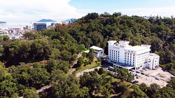 Un hotel grande y blanco con varios pisos y un patio central, rodeado de abundante vegetación y un paisaje montañoso.