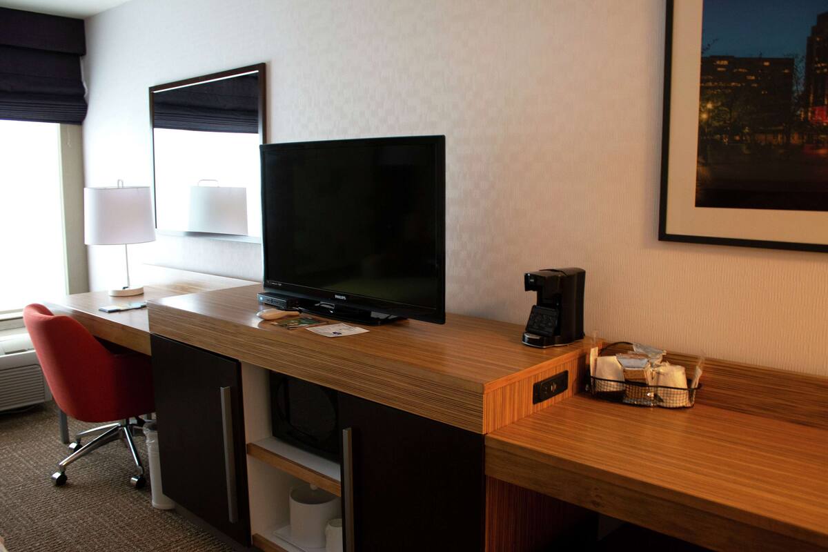 A hotel room with a wooden desk, a flat-screen TV, a red chair, a coffee maker, and a framed picture on the wall.