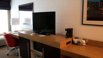 A hotel room with a wooden desk, a flat-screen TV, a red chair, a coffee maker, and a framed picture on the wall.