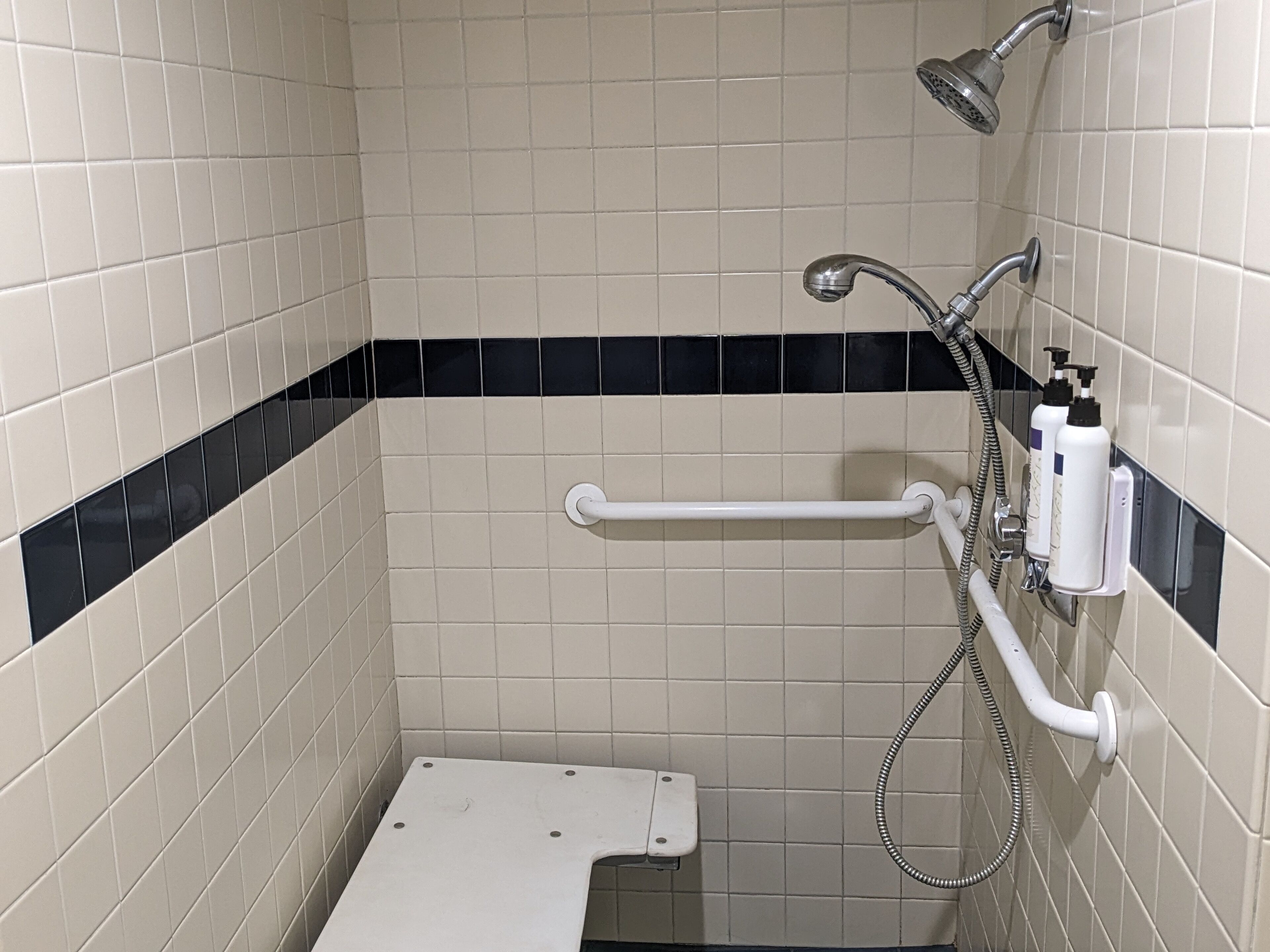 Pequeno espaço de duche revestido a azulejos, com barra de apoio, chuveiro manual e duas embalagens de champô.