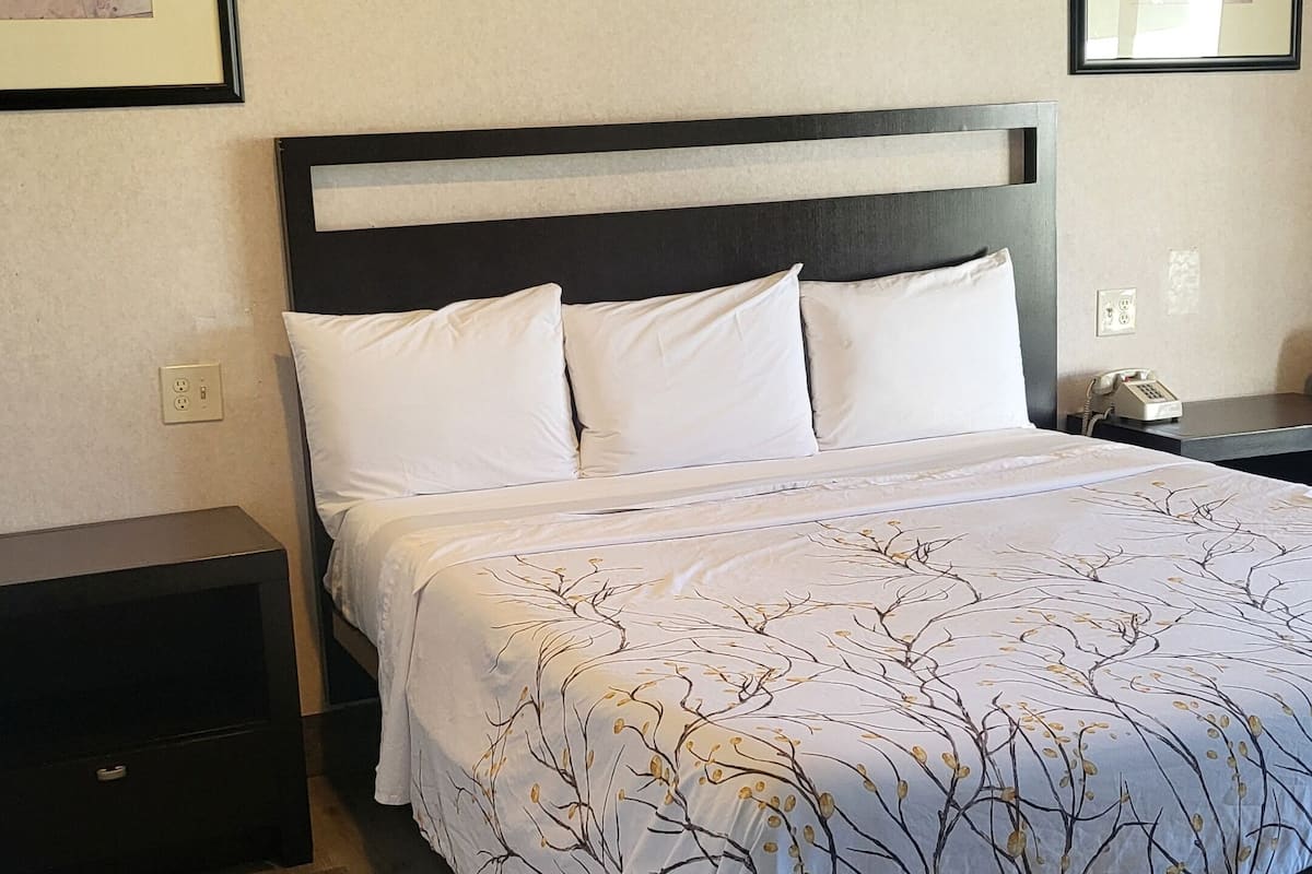 A neatly made bed with a patterned bedspread, a dark headboard, and white pillows. There are two bedside tables, one on each side of the bed. The room has wooden flooring and a textured ceiling.