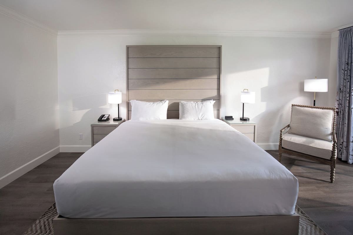 Ein Hotelzimmer mit einem großen Bett, zwei Nachttischen mit Lampen, einem Sessel und einem Fenster mit Vorhängen.