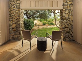 Un patio con dos sillas de mimbre, una mesa redonda pequeña, paredes de piedra y vistas a un jardín ajardinado.