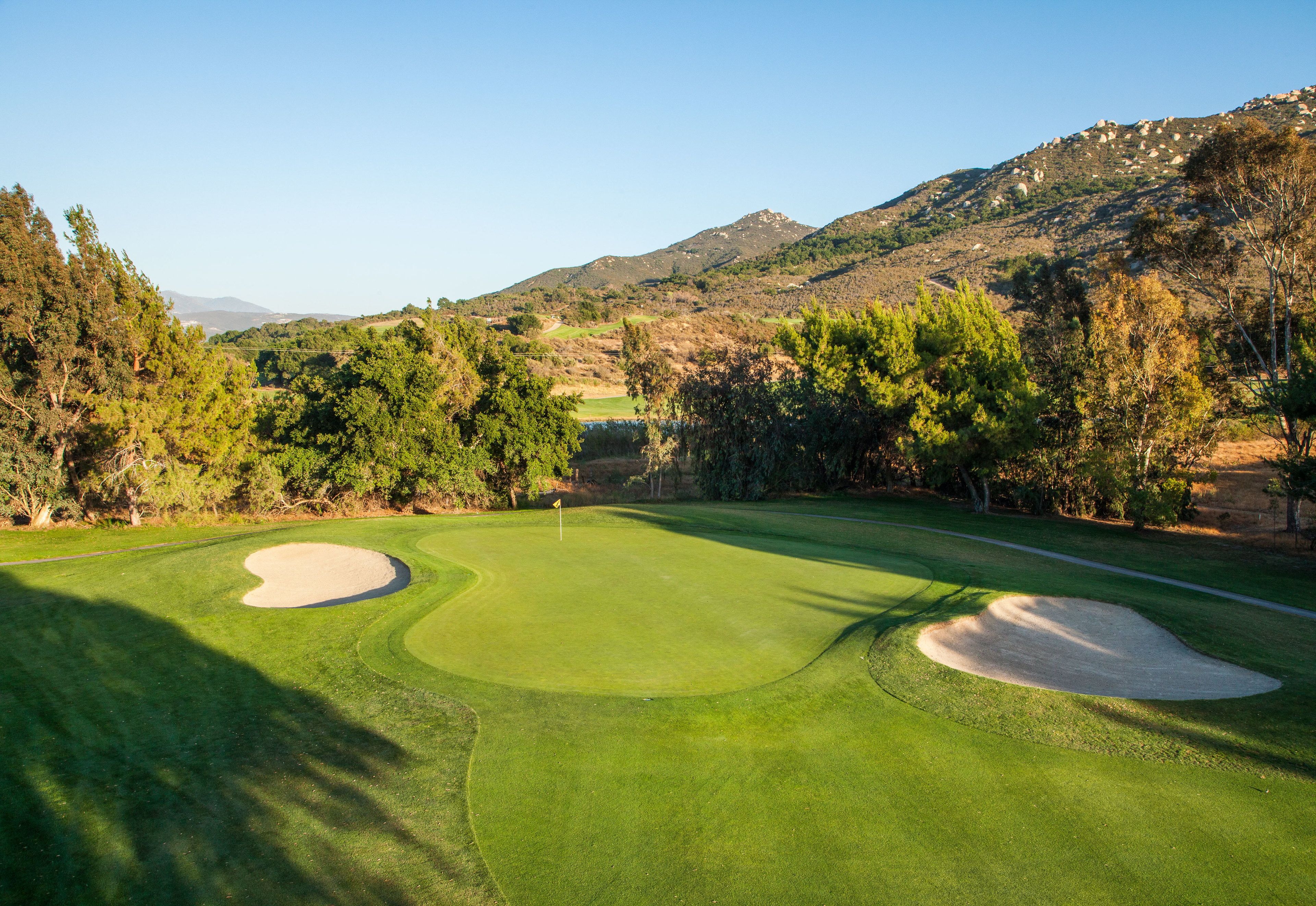 A golf course with bunkers, trees, and a mountainous backdrop.