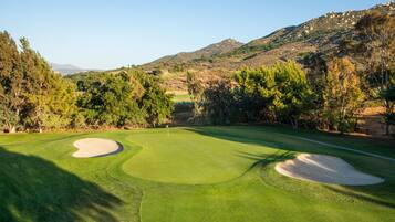 A golf course with bunkers, trees, and a mountainous backdrop.