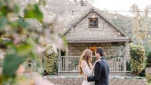 A couple embracing in front of a rustic stone building with a balcony, surrounded by greenery. - Temecula Creek Inn (Temecula)