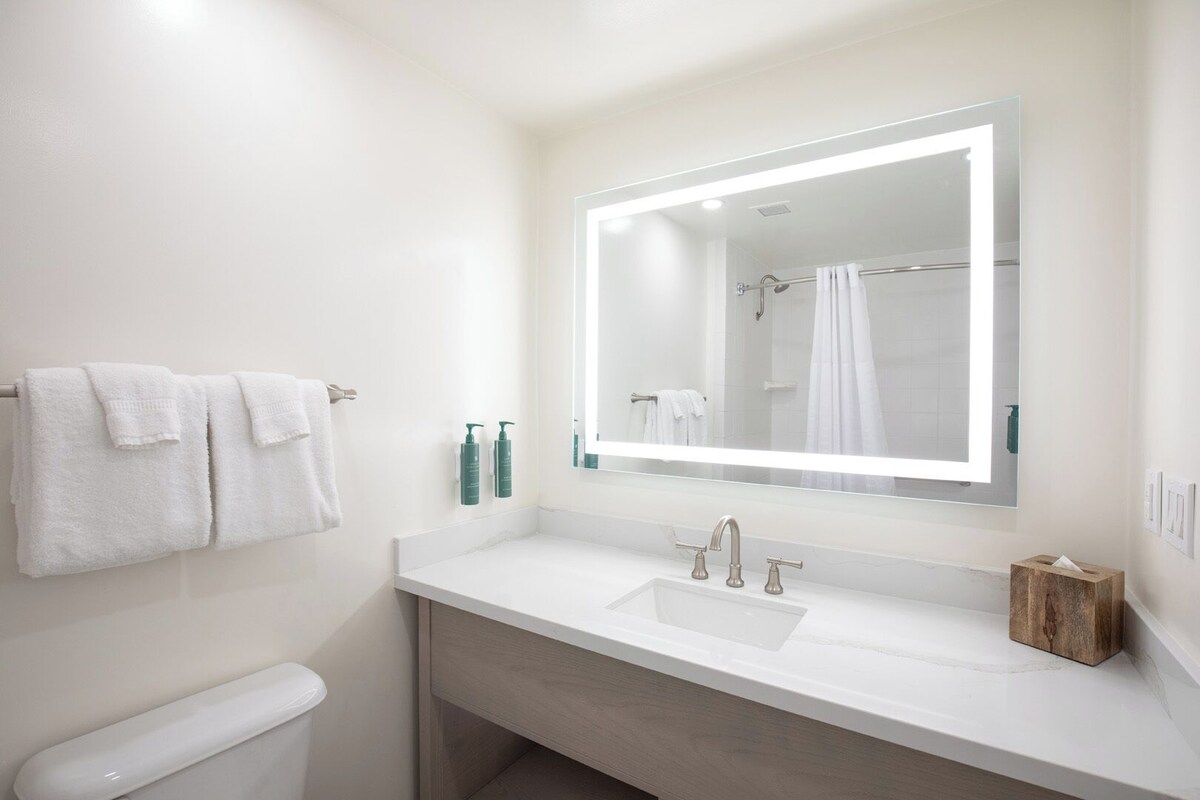 Een moderne badkamer met een wit wastafelmeubel, twee kraanwerkstukken, een spiegel met ingebouwde verlichting en een handdoekrek met twee witte handdoeken.