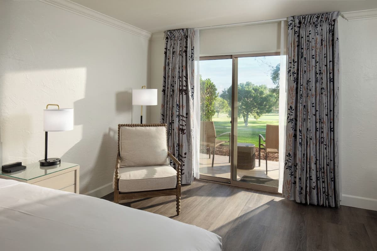 Ein Hotelzimmer mit Bett, Sessel, Lampe und Blick auf einen Golfplatz durch eine Schiebetür.