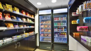 Zona de snacks y bebidas con estantes de papas fritas, un refrigerador con bebidas y una pared con artículos colgantes.