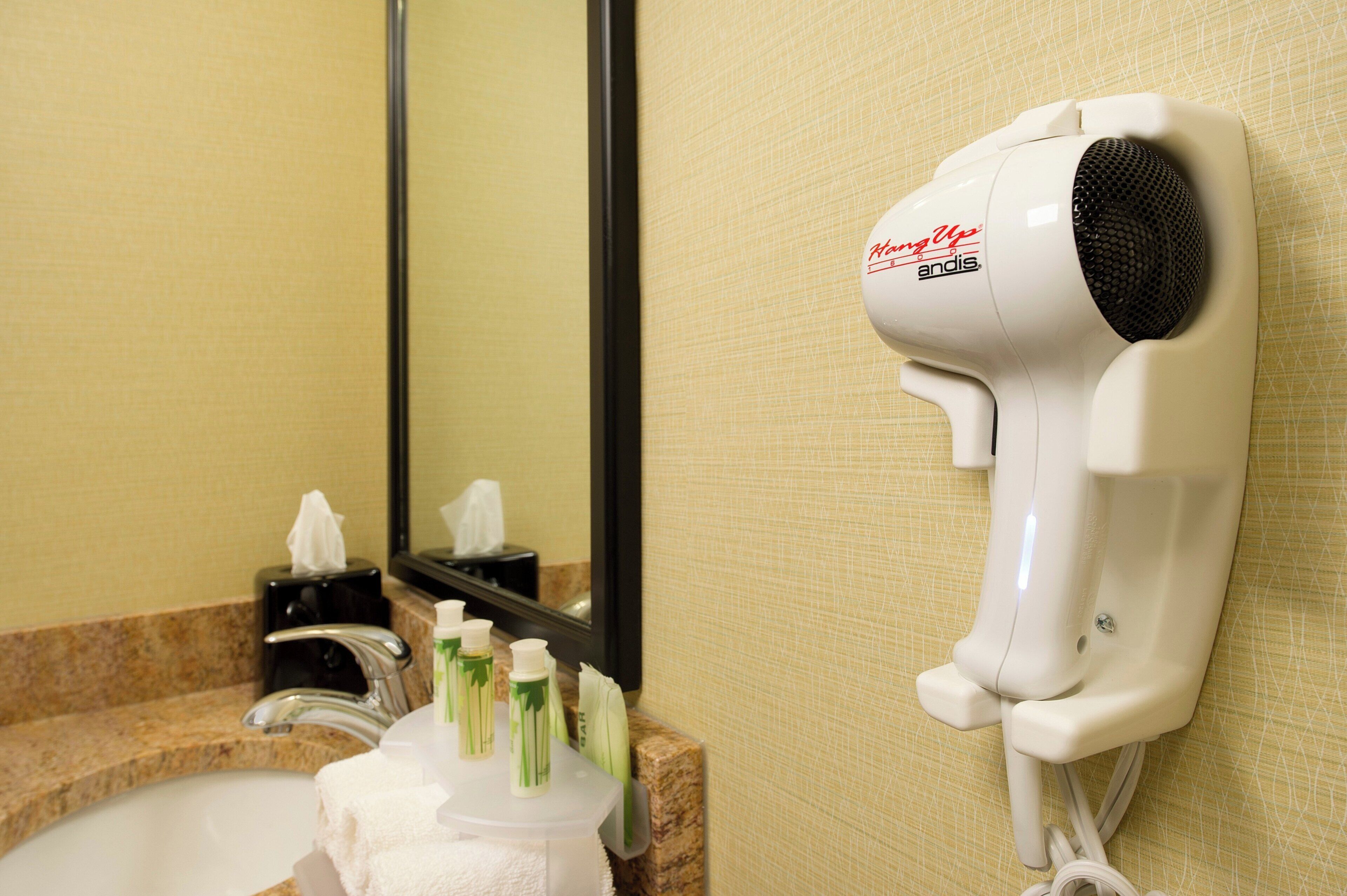 A hair dryer mounted on a wall in a bathroom with a mirror, a sink, and toiletries.