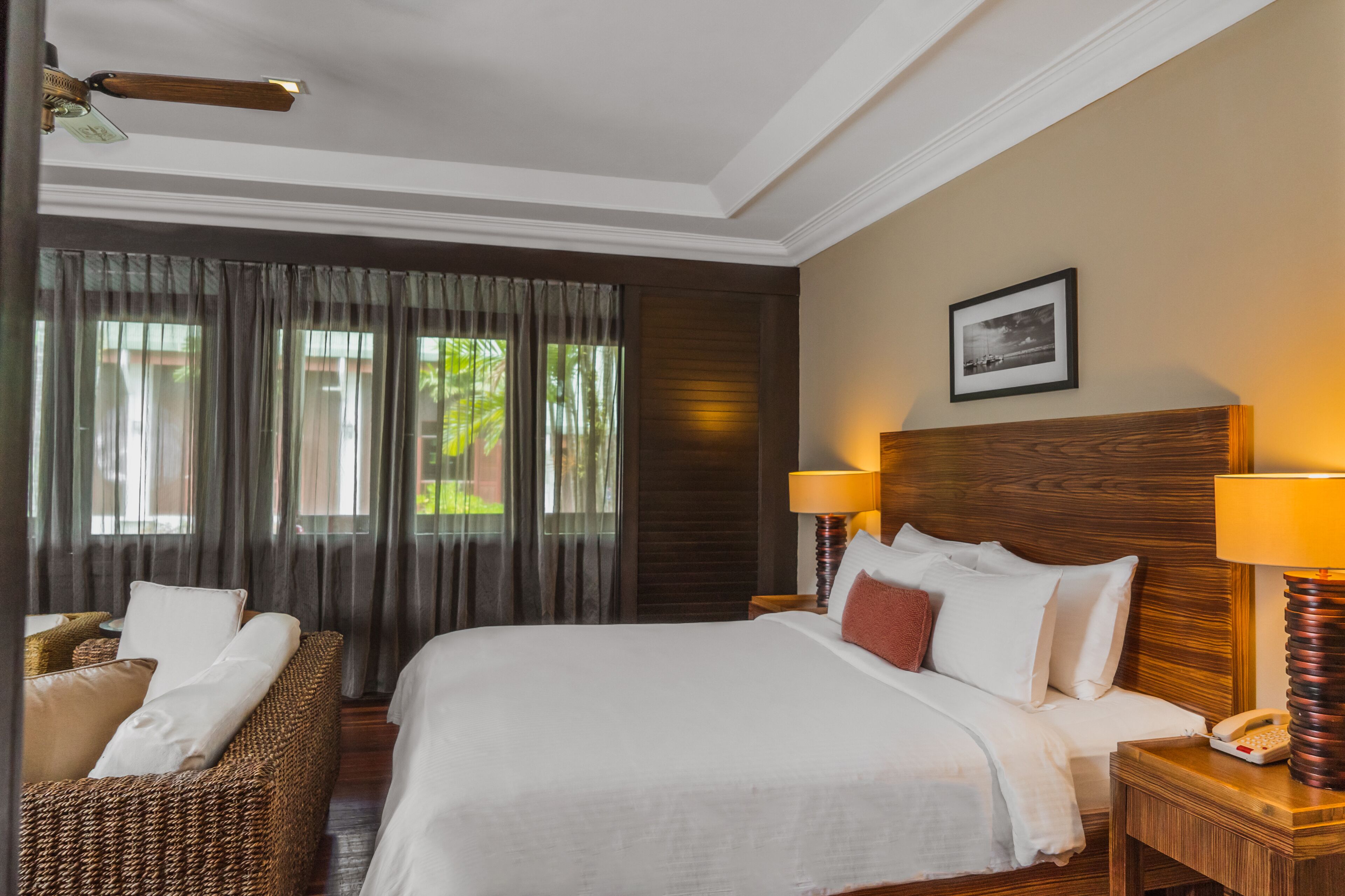 Habitación de hotel con una cama grande, dos lámparas de noche, un cabecero de madera, un cuadro enmarcado en la pared y una ventana con cortinas.