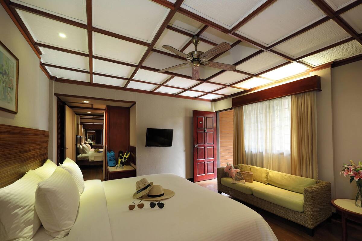A hotel room with a large bed, a sofa, a television, a ceiling fan, and a window with curtains.