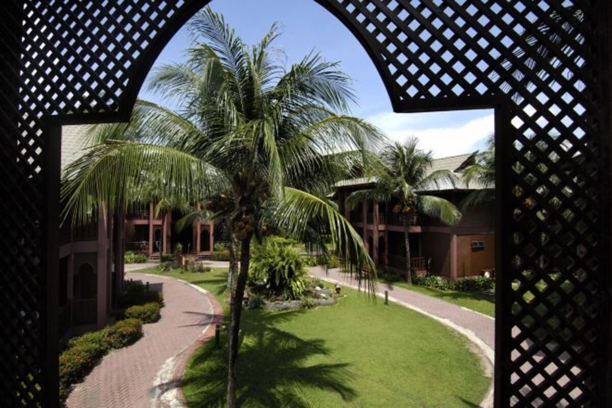 A view through a lattice structure, featuring a pathway, palm trees, and a building with a modern architectural style.
