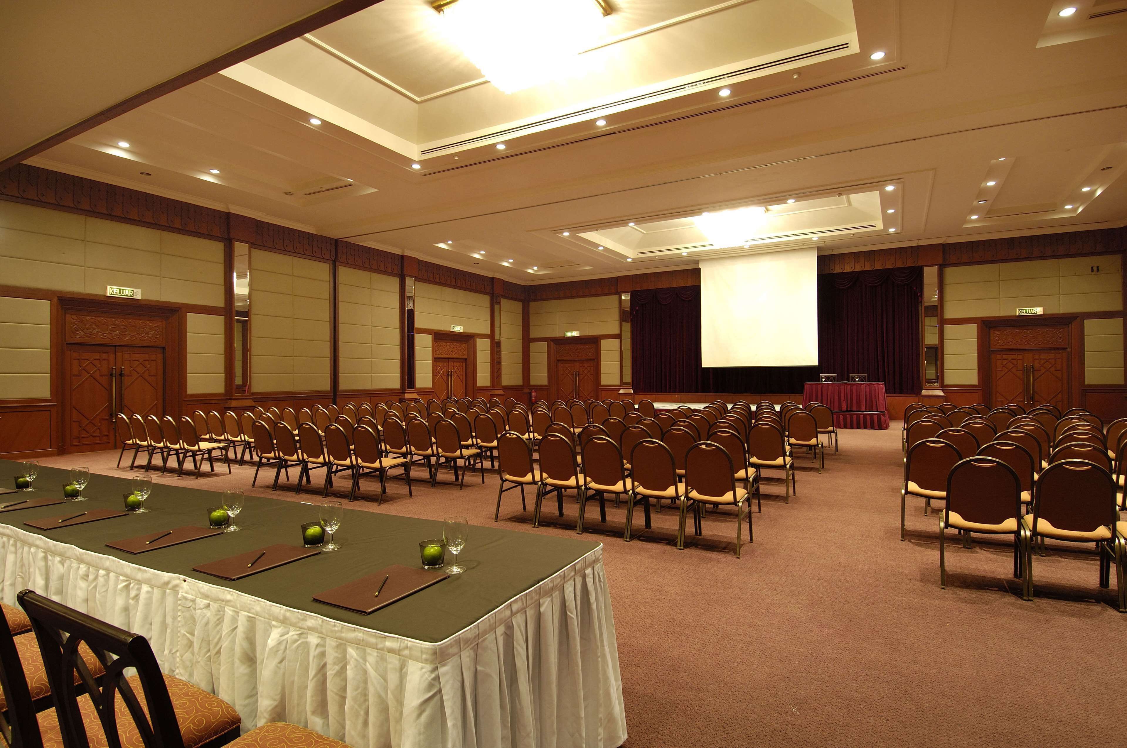A conference room with rows of chairs, a long table with place settings, and a stage with a screen.