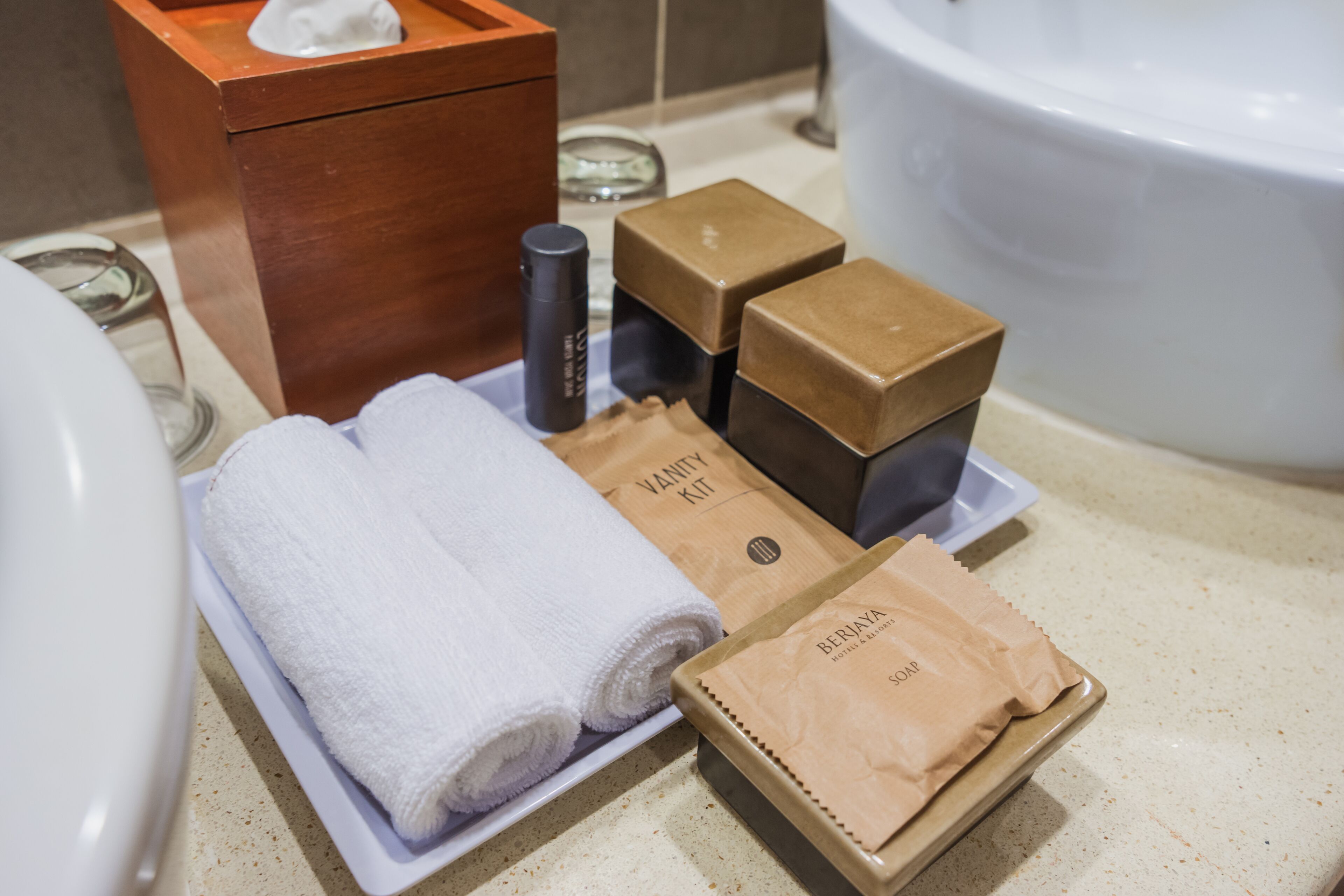 Un tocador de baño con un lavabo blanco, un mueble de madera y una bandeja con artículos de aseo que incluye una toalla blanca, una botella negra y dos cajas marrones con la etiqueta "BERIYA".