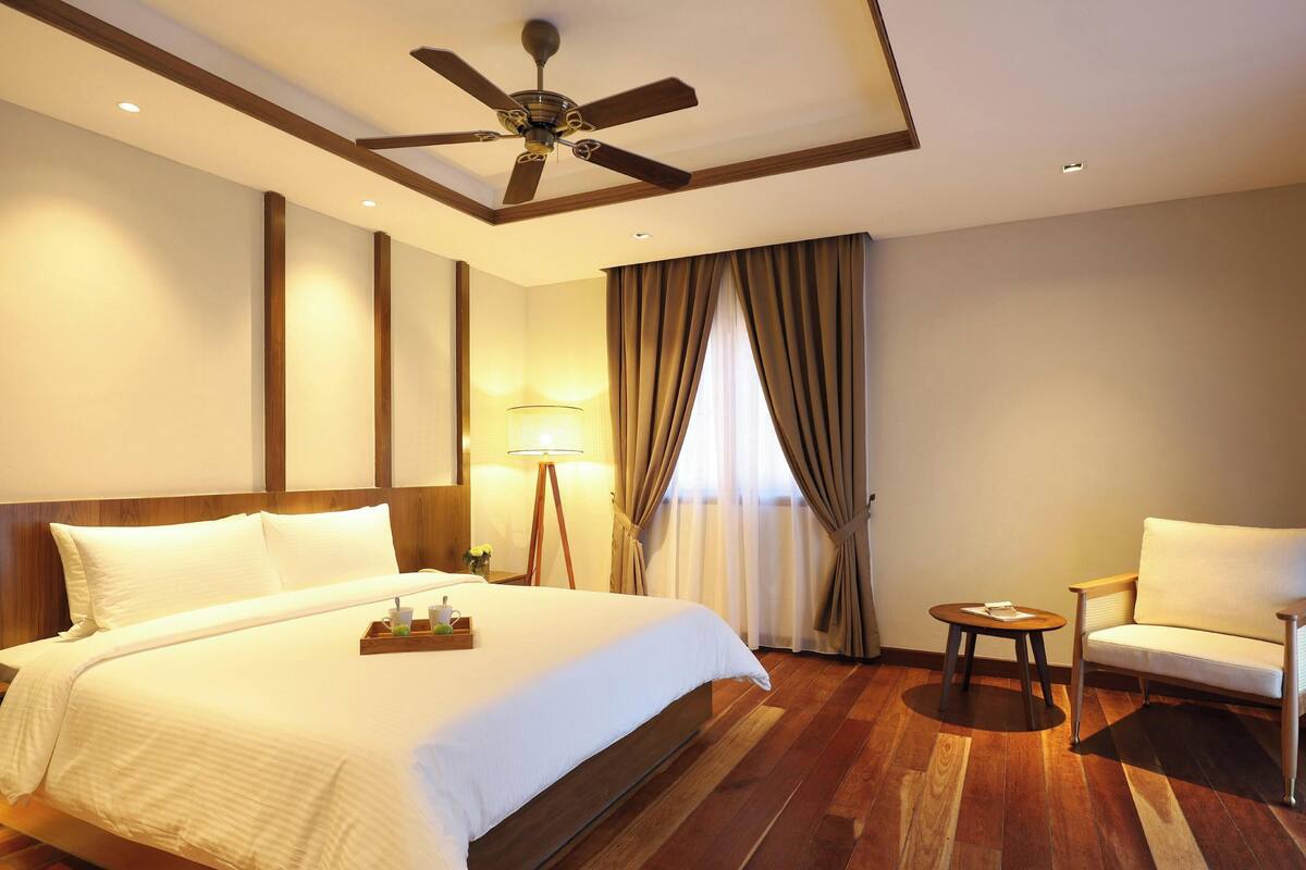 A hotel room with a large bed, a ceiling fan, a small table, a chair, and a lamp.