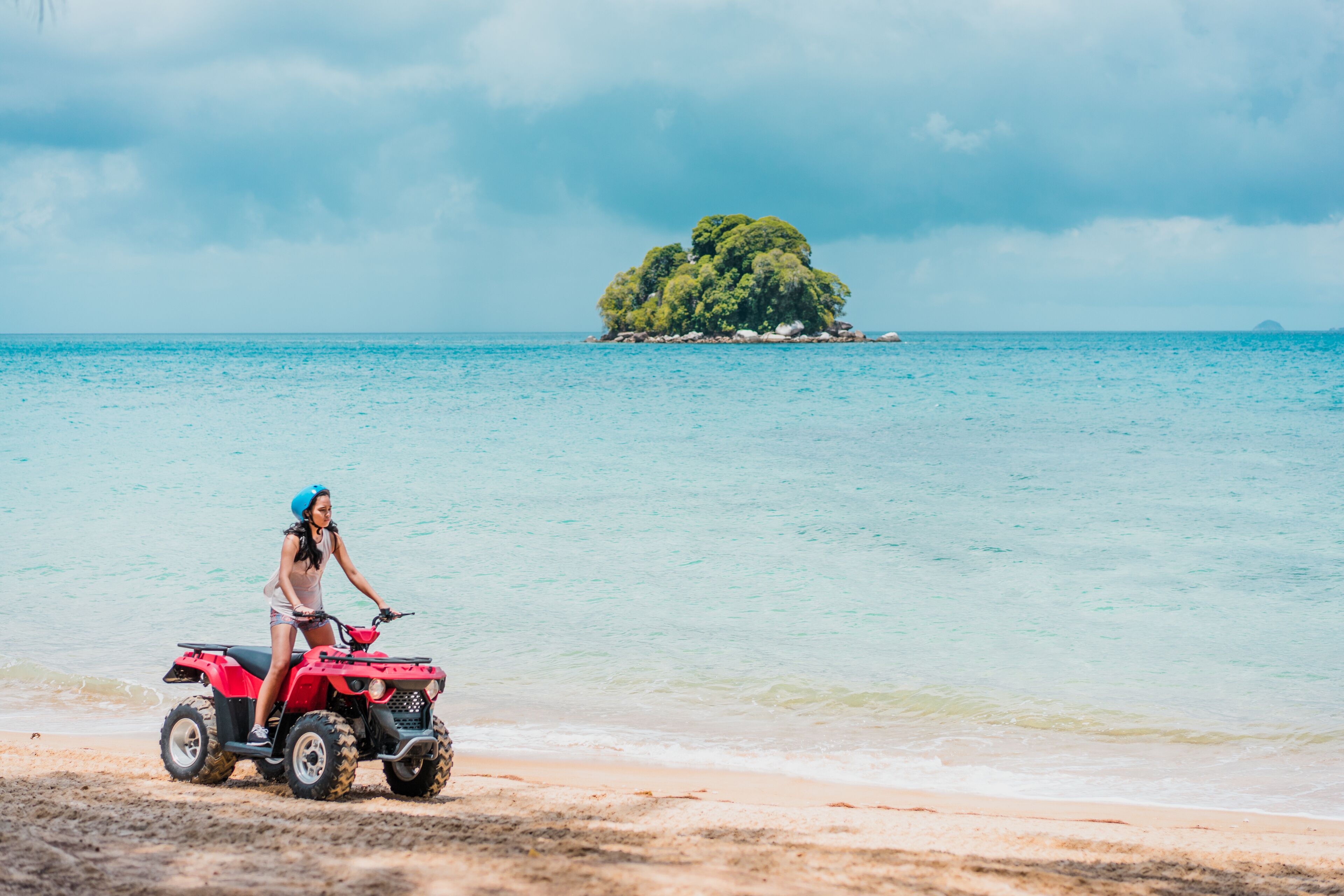 A person on a red ATV on a sandy beach with clear turquoise water and a small island in the distance.