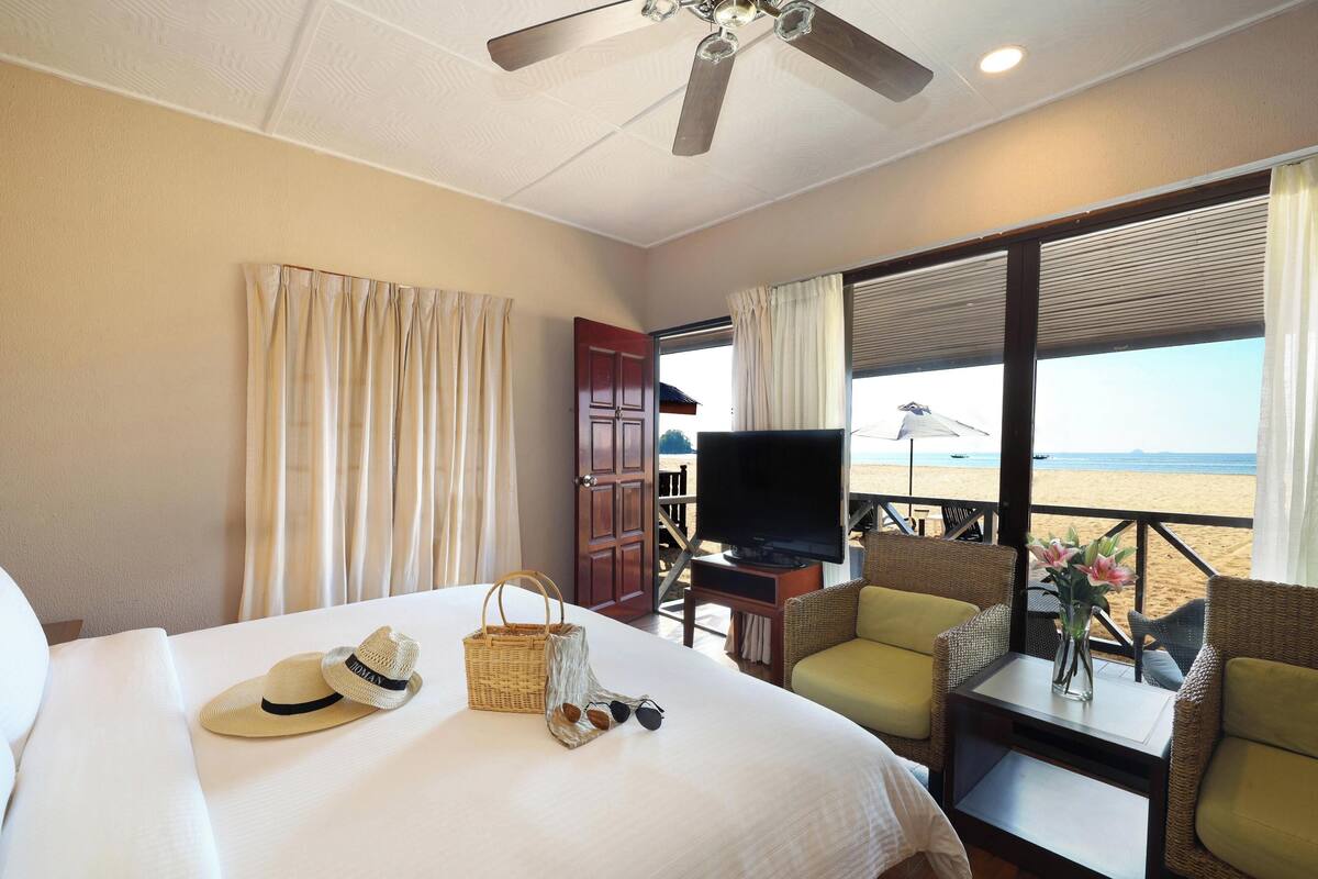 A hotel room with a bed, a television, wicker furniture, and a view of the beach.