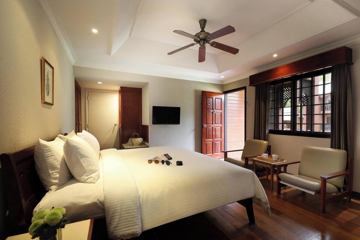 A hotel room with a large bed, a ceiling fan, a TV, a window with a view, and a small table with a chair.