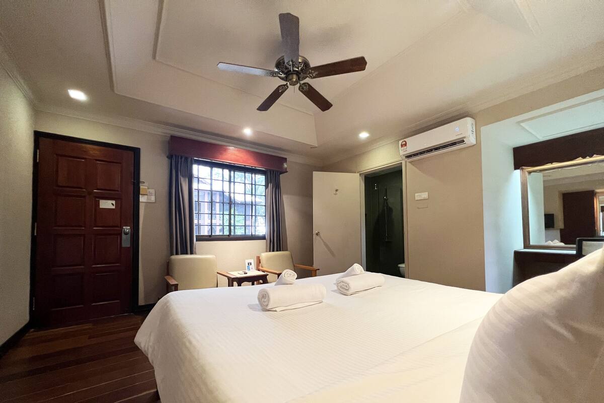 A hotel room with a bed, two chairs, a desk, a ceiling fan, a window with curtains, an air conditioning unit, and a door.