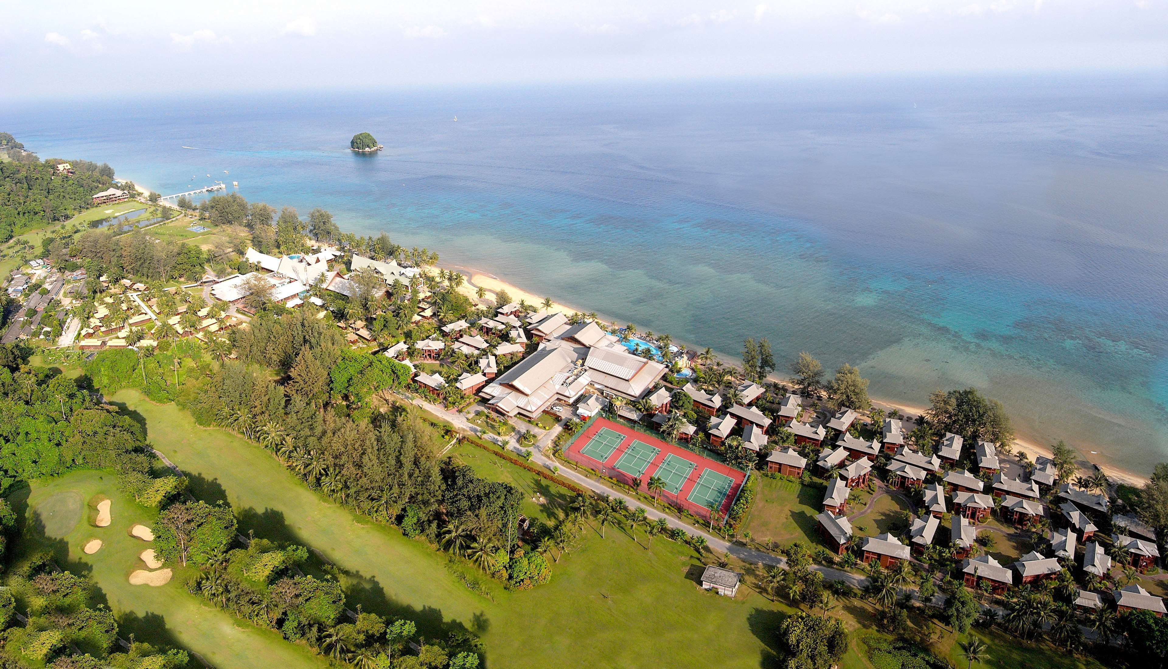 Vue aérienne d’une station balnéaire comprenant de nombreux hébergements de type bungalow, un bâtiment central doté d’un toit rouge, et offrant une vue dégagée sur l’océan et une petite île.