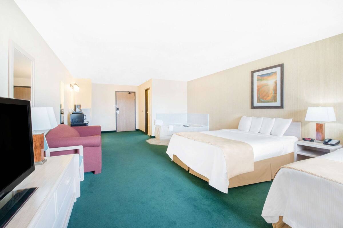 Een hotelkamer met een bed, een bureau met een computer, een stoel, een televisie, een nachtkastje met een lamp, en een schilderij aan de muur.