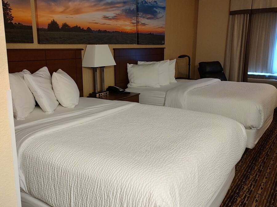 Quarto de hotel com duas camas, uma mesinha de cabeceira, um abajur, uma cadeira e um quadro grande de um pôr do sol.